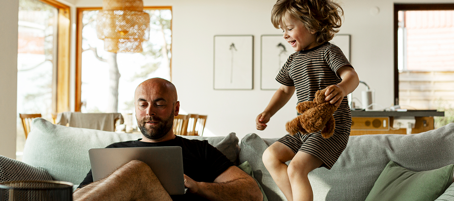 Father sitting on a sofa using a laptop and child holding a teddy and jumping next to him